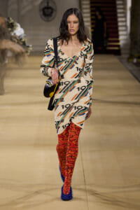 Model walking the runway in a loose, patterned dress with bold orange, teal, and black numbers; red swirl tights and blue furry heels, carrying a black shoulder bag with yellow trim.