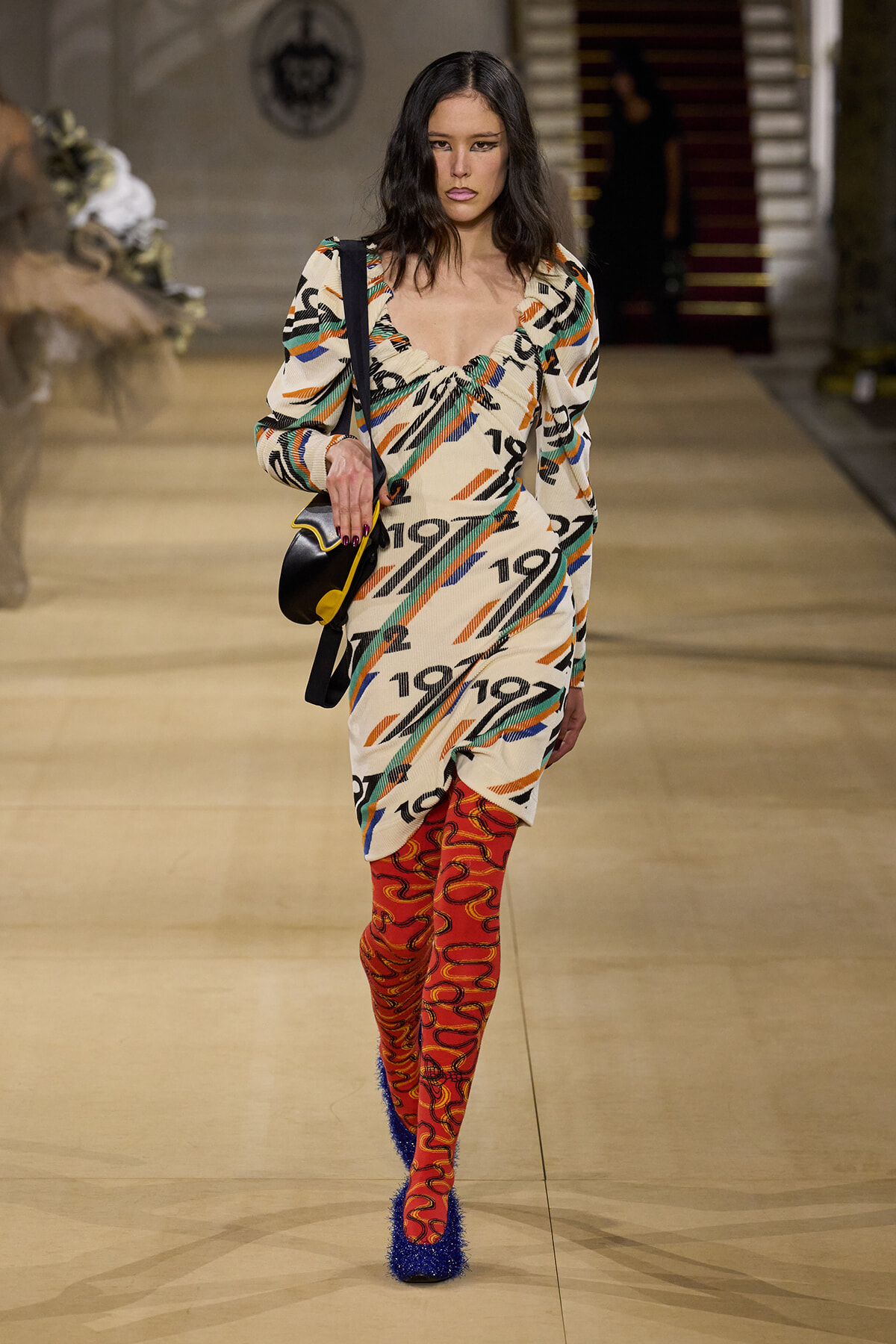 Model walking the runway in a loose, patterned dress with bold orange, teal, and black numbers; red swirl tights and blue furry heels, carrying a black shoulder bag with yellow trim.
