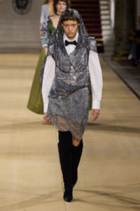 Runway model in a silver sequin hooded top over a white shirt with a black bow tie, metallic mesh skirt, and black knee-high boots.