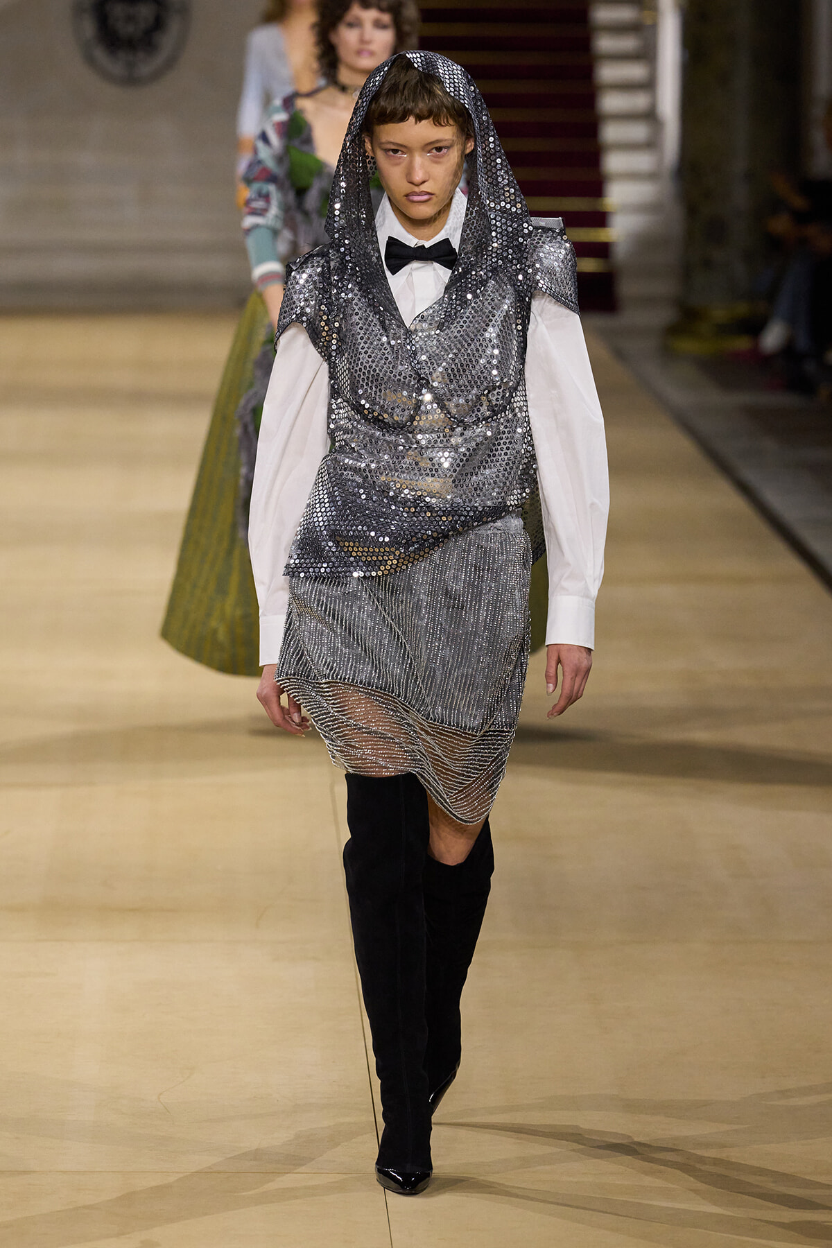 Runway model in a silver sequin hooded top over a white shirt with a black bow tie, metallic mesh skirt, and black knee-high boots.