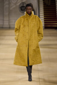 Model on a fashion runway wearing a long, oversized mustard faux‑fur coat with hands in pockets and bold dangling earrings.