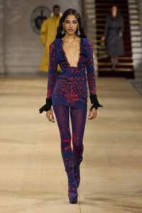 Runway model wearing a blue and red floral knit dress with a deep plunging neckline, ruffled shoulders, and matching purple tights and boots.