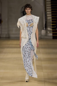 Model on a runway wearing a cream crocheted cardigan over a pale blue polka-dot dress with lace panels and knee-high boots.