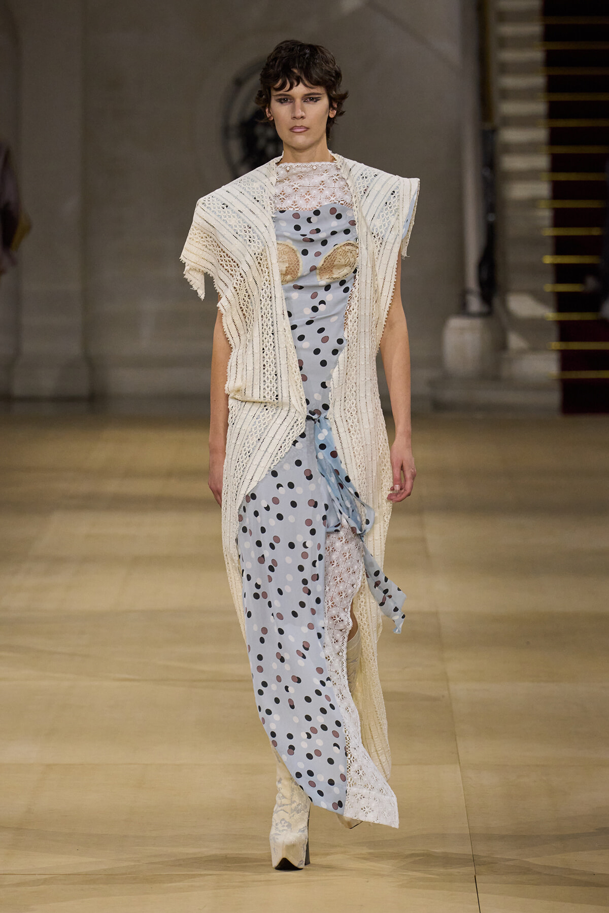 Model on a runway wearing a cream crocheted cardigan over a pale blue polka-dot dress with lace panels and knee-high boots.