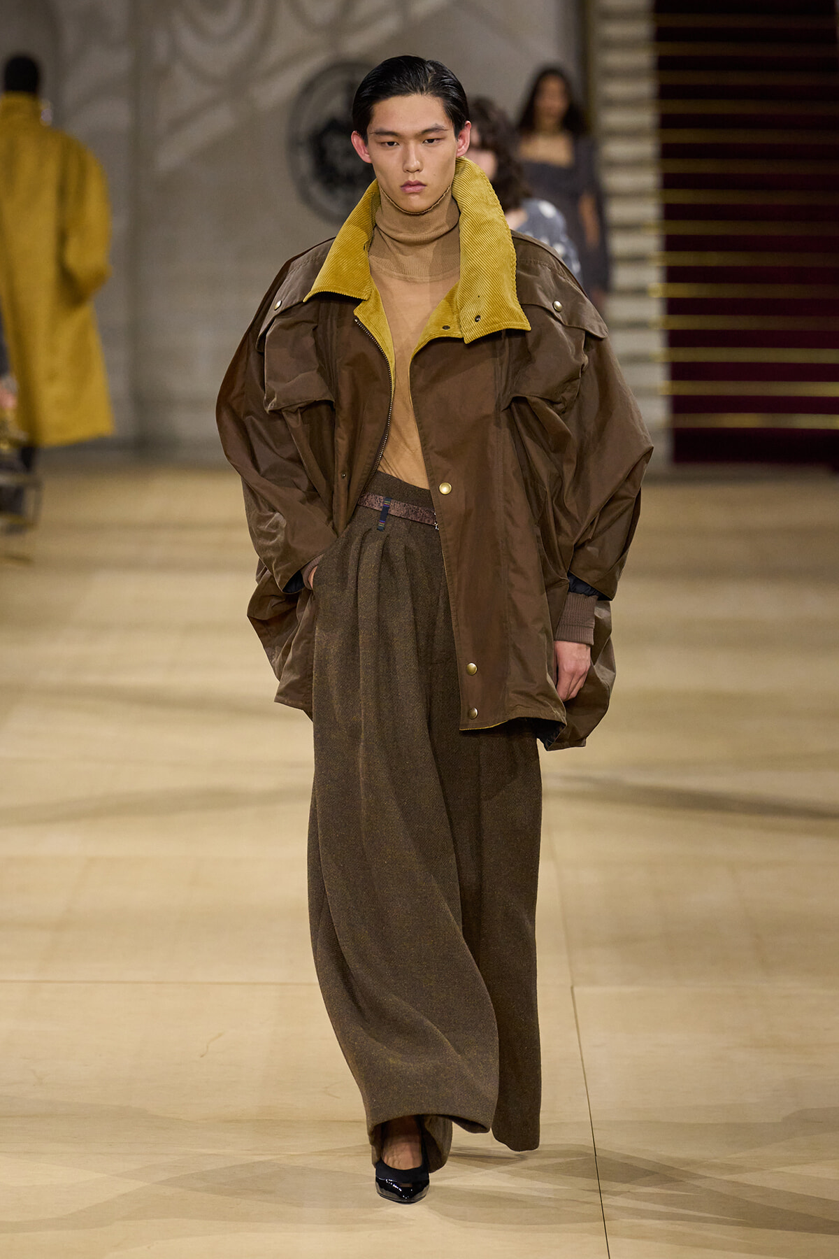 Model walks the runway in an oversized brown jacket with a mustard-yellow collar, beige turtleneck, and wide brown trousers with a brown belt, finishing with black heels.