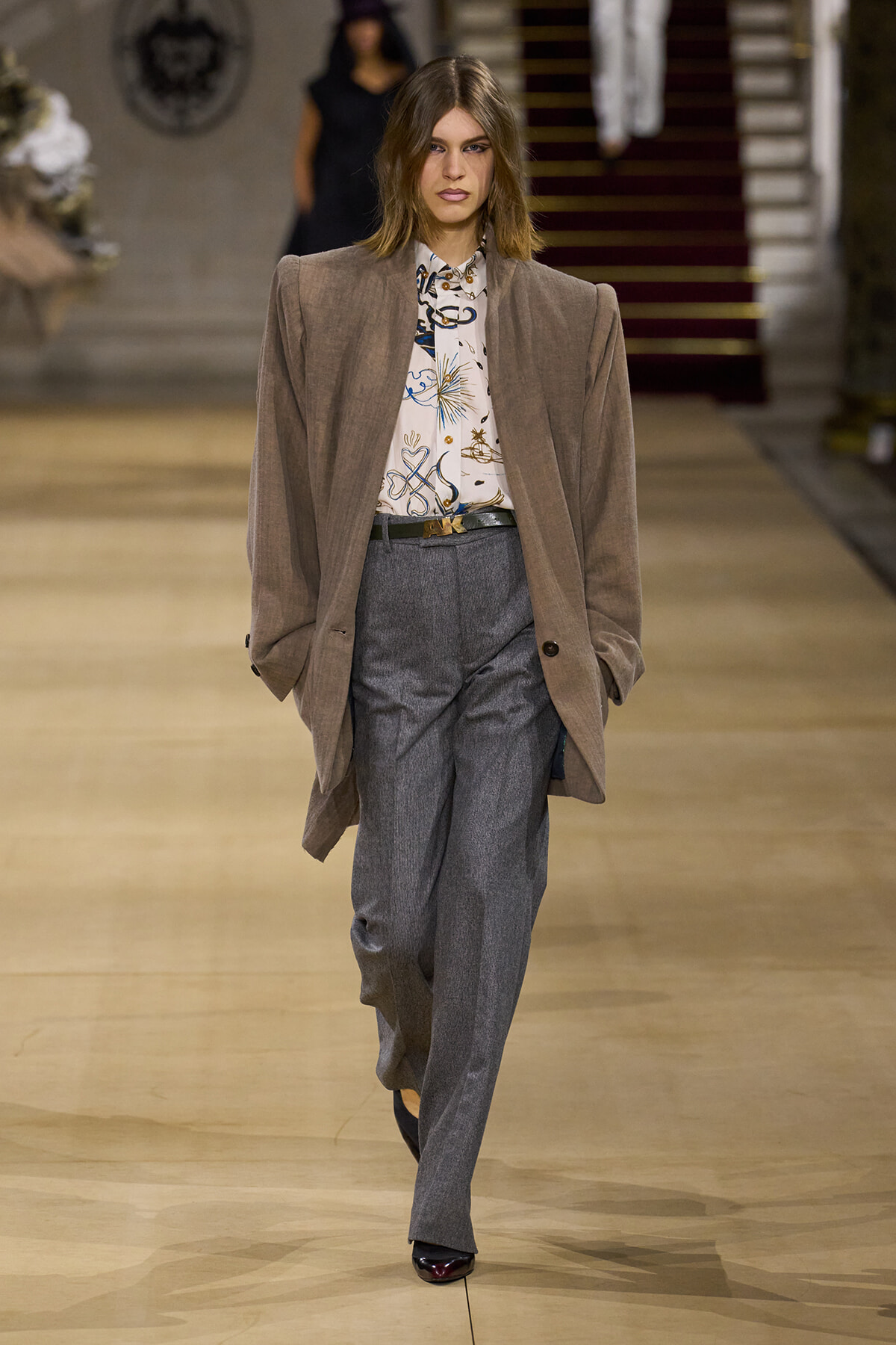 Model walking a fashion show runway in an oversized taupe blazer, patterned blouse, and high-waisted grey trousers with a black belt and dark loafers.
