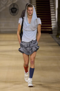 Male model walks a fashion runway wearing a light patterned hooded top with a loose v-neck, gray patterned shorts, mismatched socks (red and blue), and white high-top sneakers, carrying a black bag over a shoulder.