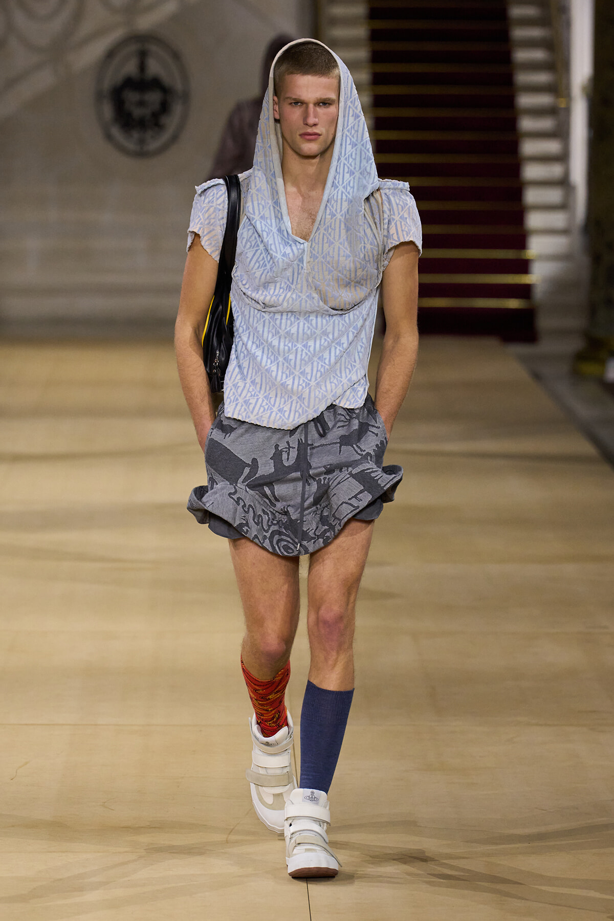 Male model walks a fashion runway wearing a light patterned hooded top with a loose v-neck, gray patterned shorts, mismatched socks (red and blue), and white high-top sneakers, carrying a black bag over a shoulder.