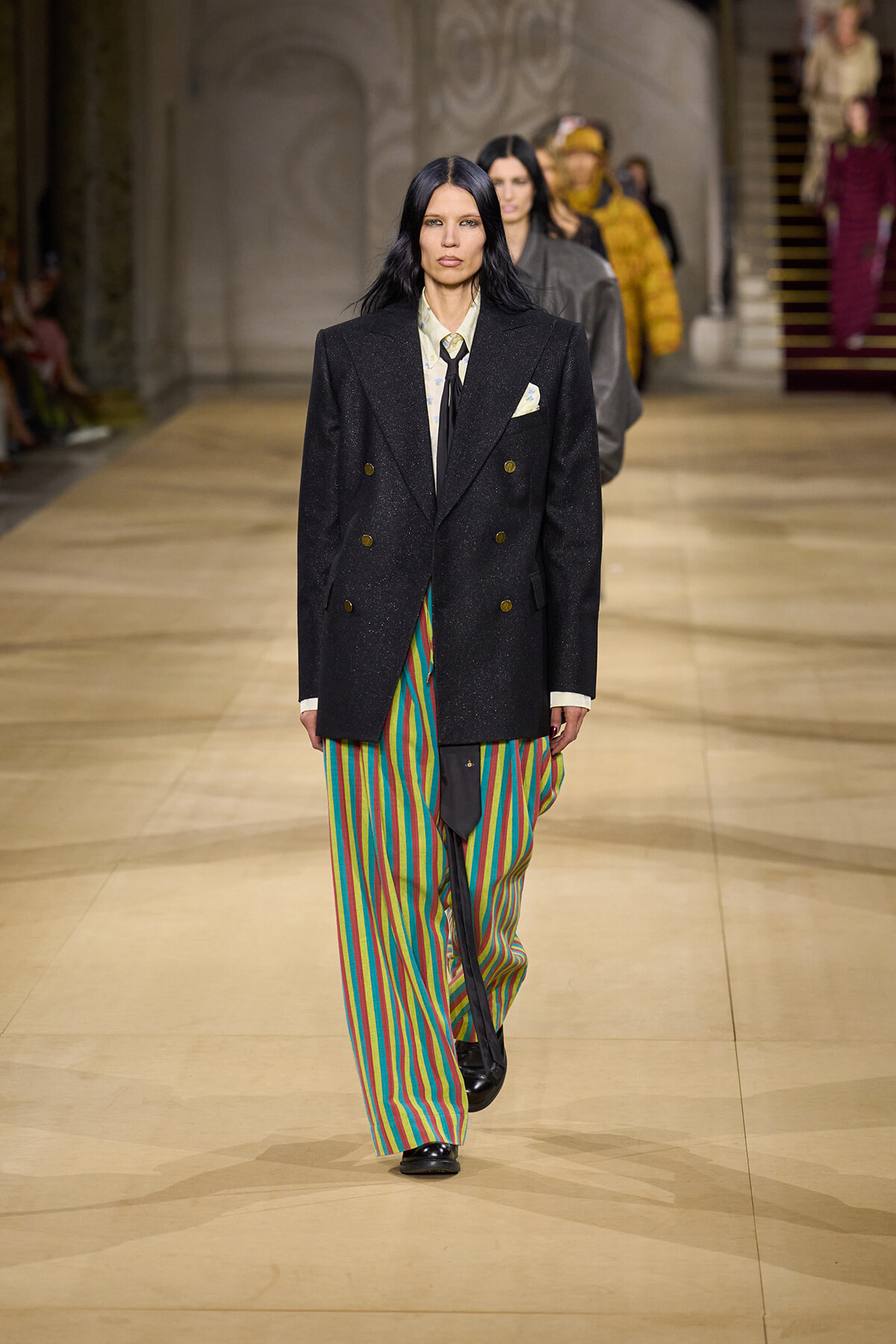Model walking a runway in a black double-breasted blazer, white shirt, tie, and bright striped pants.