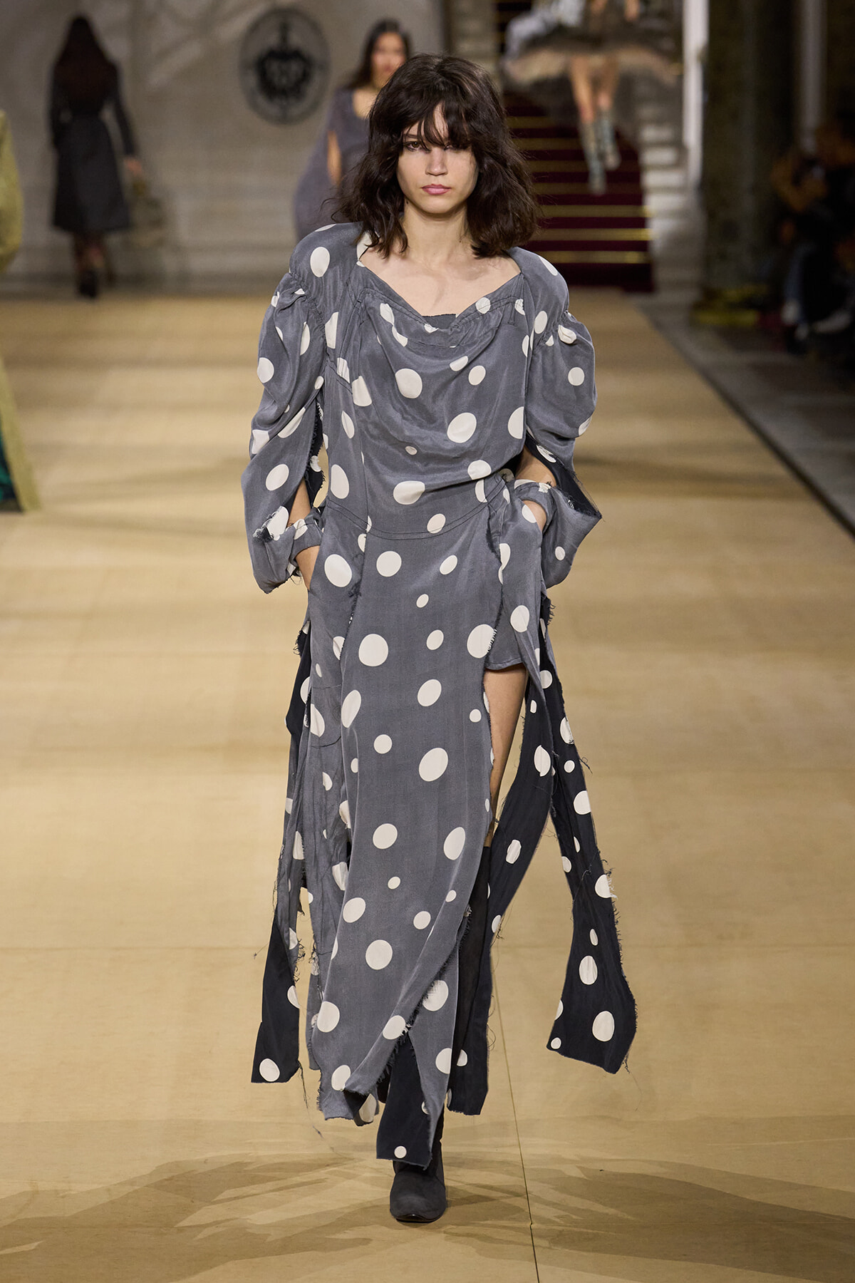 Model walking a fashion show in a grey polka-dot maxi dress with voluminous sleeves and a front slit in the skirt, neutral makeup.