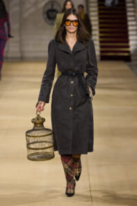 Model walking a fashion runway in a dark gray trench coat with a belted waist, oversized collar, and gold buttons, wearing orange sunglasses and plaid boots while holding a decorative birdcage.