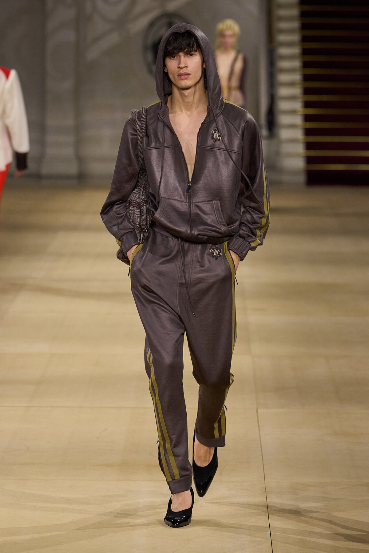 Male model in a dark gray hooded tracksuit with yellow side stripes, hands in pockets, walking the runway in black heels.