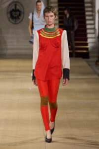 Runway model wearing a bright red tunic with gold arc detailing over white sleeves, red leggings, and brown knee pads, pairing with black heels.