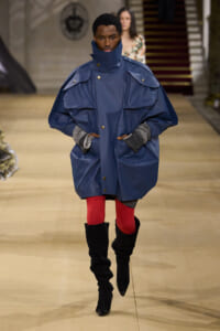 Model walks the runway in an oversized navy blue coat with a high collar, paired with red tights and black knee-high boots.
