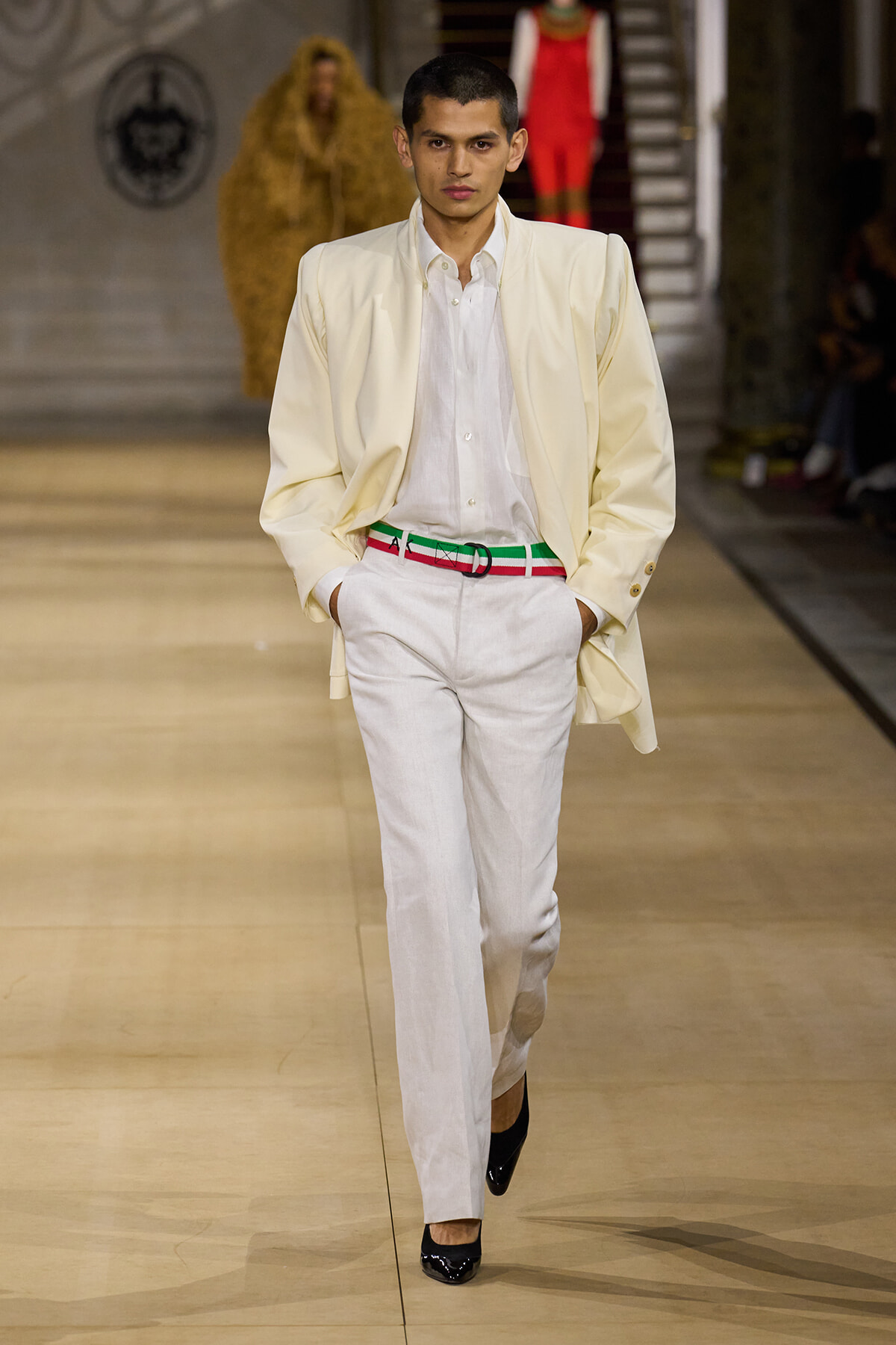 Male model walking a fashion runway in a cream suit, white shirt, and a green-red belt, hands in pockets.