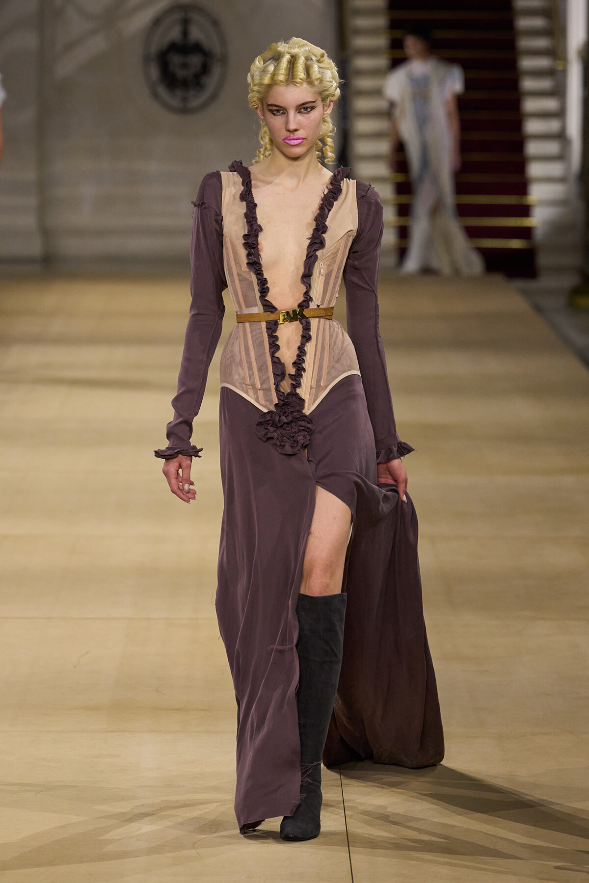 Model walking a runway in a burgundy-purple, Victorian-inspired gown with a corseted beige bodice, ruffles, belt, and black knee-high boots.