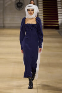 Female model walks a runway in a navy off-the-shoulder gown with a gathered neckline, black lace-up boots, and a white head wrap.