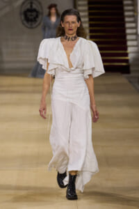 Model walks the runway in a white flowing dress with dramatic ruffled cape sleeves and a bold 'CHAOS' choker, black boots visible at the hem.