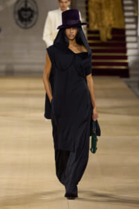 Model on a runway wearing a black oversized hooded dress with a purple wide-brim hat and a green handbag.