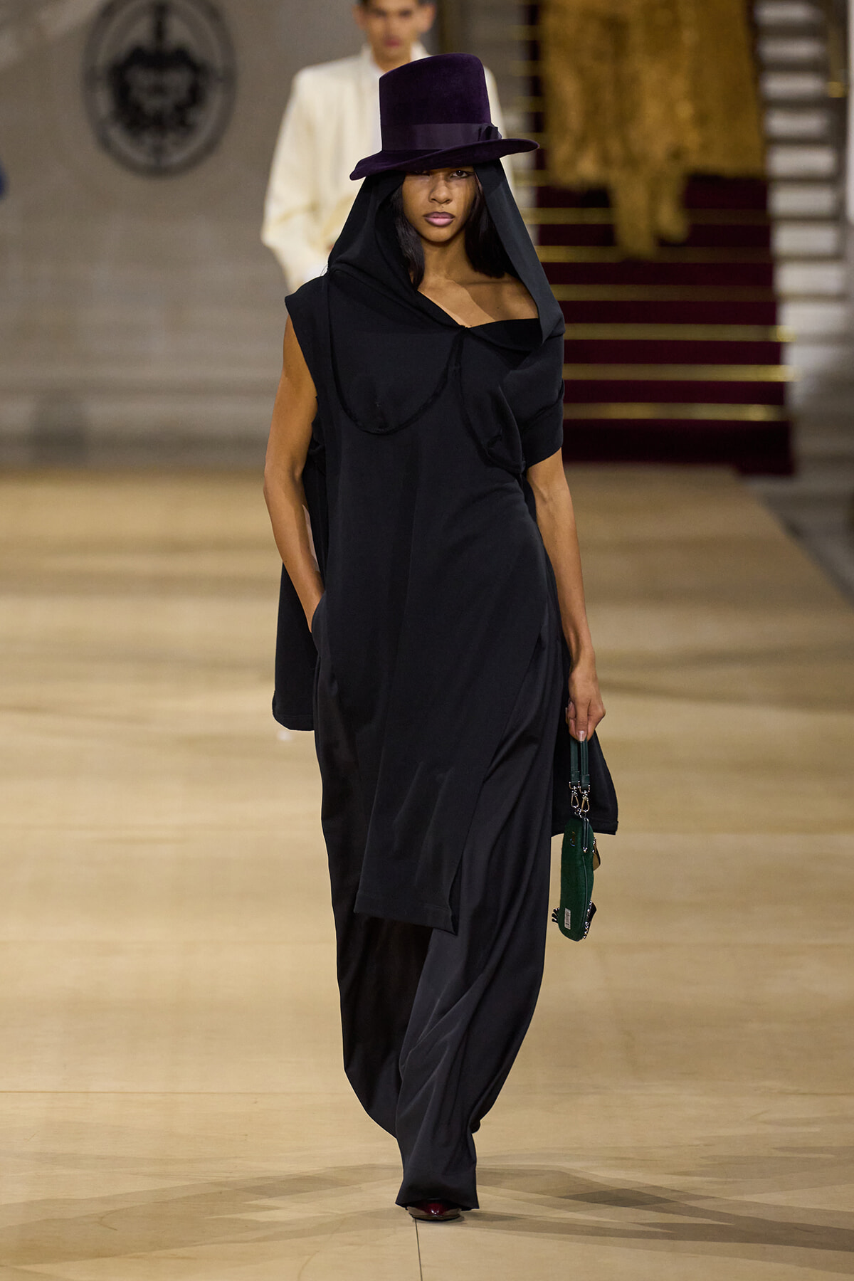 Model on a runway wearing a black oversized hooded dress with a purple wide-brim hat and a green handbag.