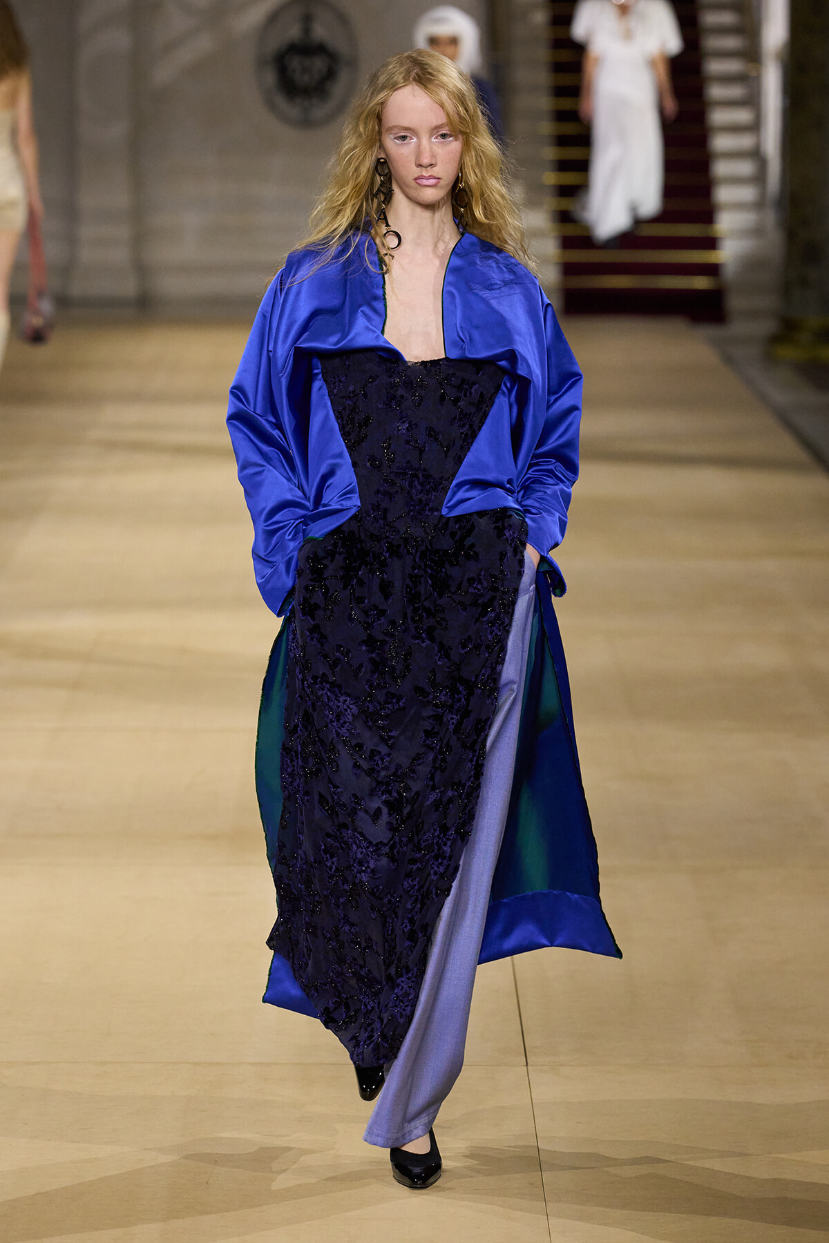 Model walking a runway in a royal blue satin overcoat over a black lace dress with teal panels showing at the sides.