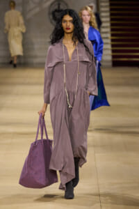 Model walking the runway in a dusty mauve maxi dress with a plunging neckline, layered pearl necklace, and a large purple tote bag.