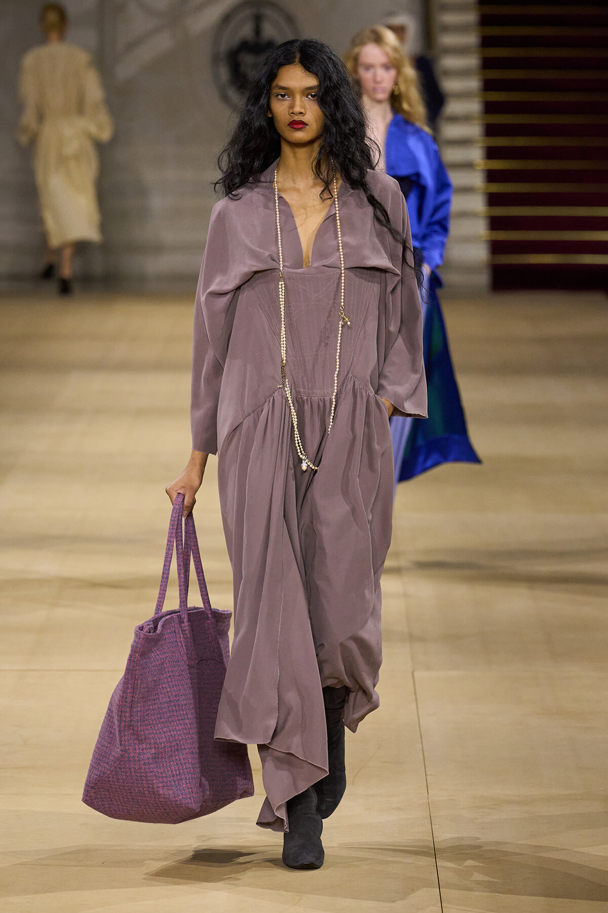 Model walking the runway in a dusty mauve maxi dress with a plunging neckline, layered pearl necklace, and a large purple tote bag.