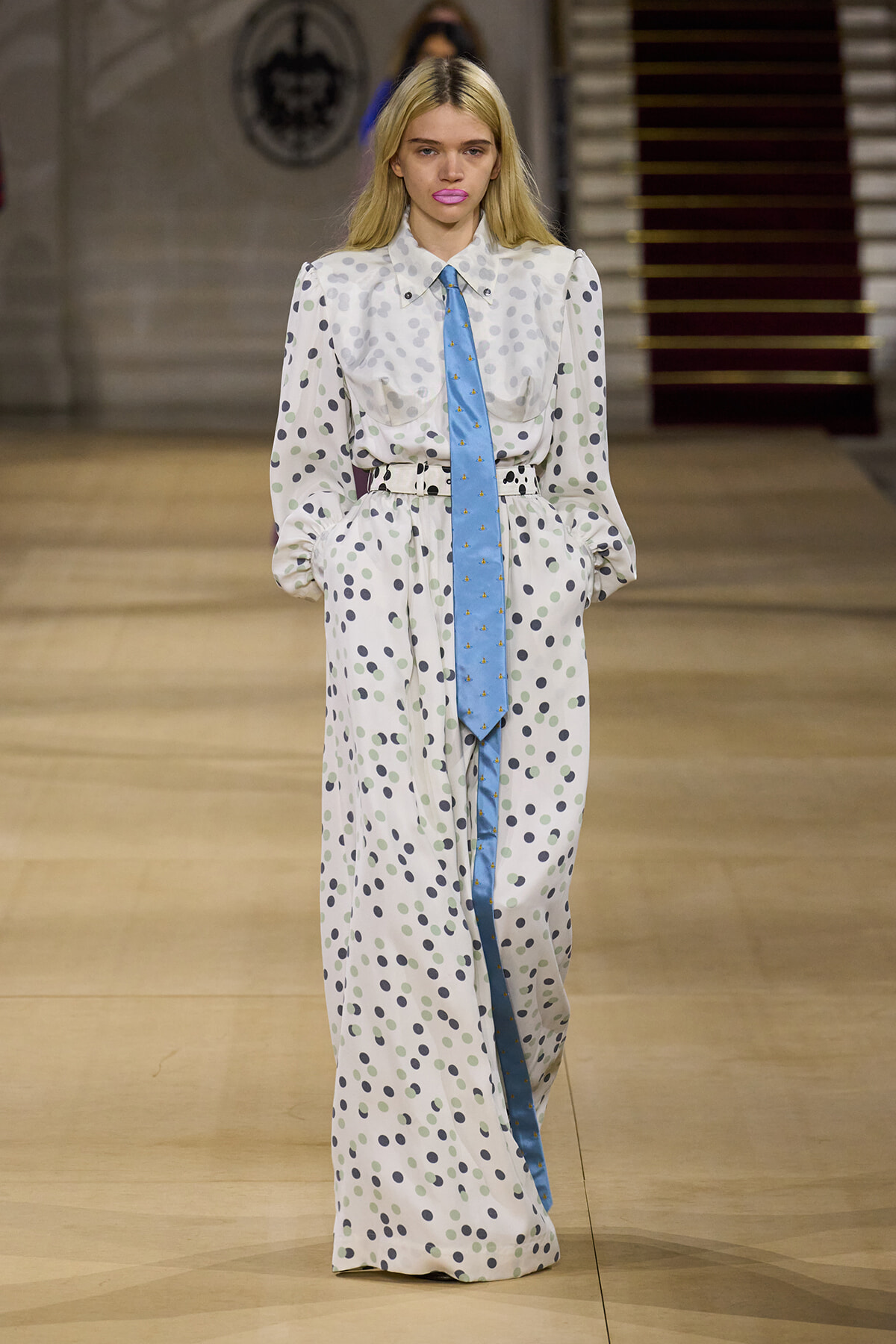 Runway model wearing a long white polka-dot dress with a light blue necktie and belt, walking on a fashion show runway.
