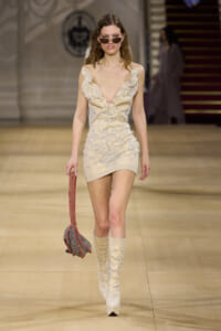 Model walking a runway in a cream floral mini dress with matching knee-high boots, sunglasses, and a red-striped handbag.