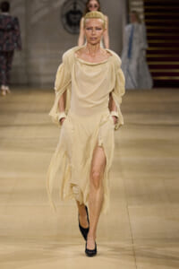 Model walking a beige flowing dress with a high slit on a fashion runway, wearing black heels and her hair pulled back
