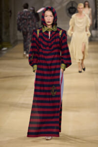 Model walks the runway in a long red-and-navy striped dress with an anchored embroidery, green collar, and hooded headpiece.