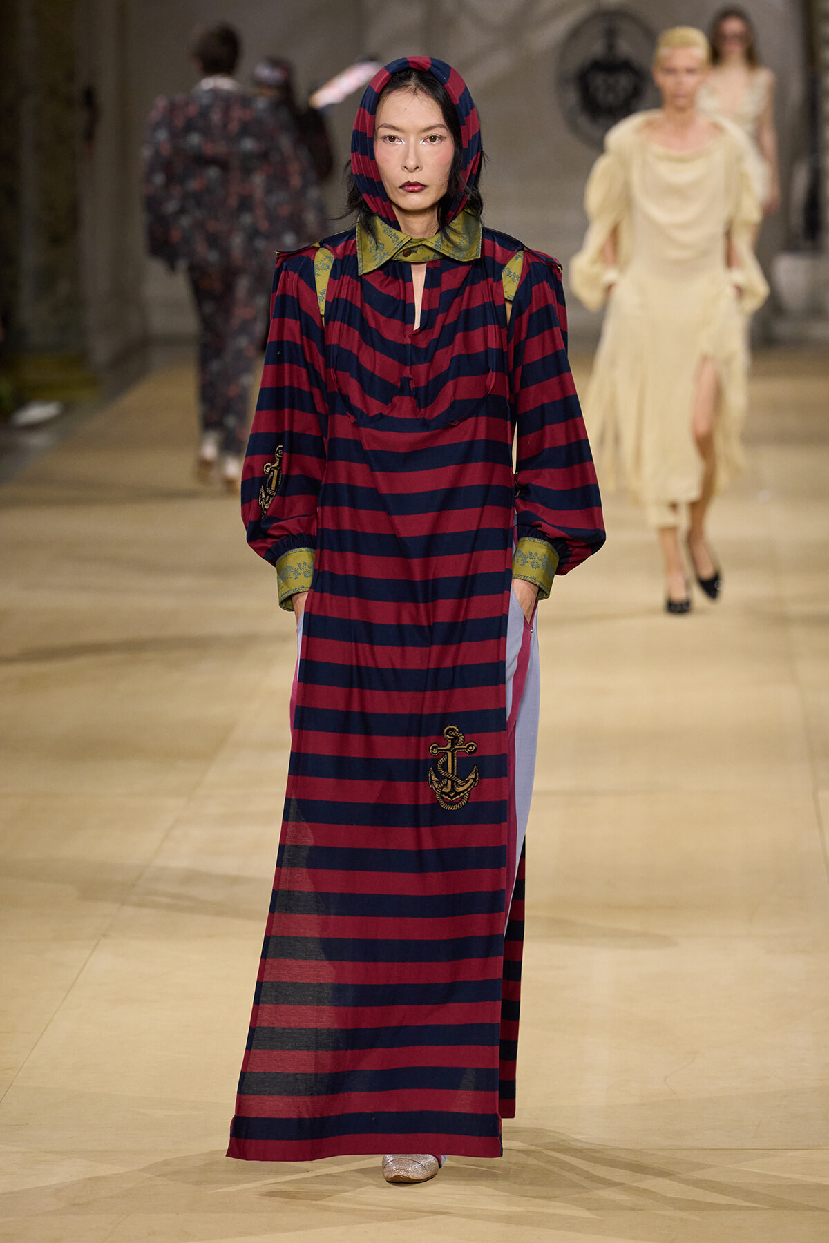 Model walks the runway in a long red-and-navy striped dress with an anchored embroidery, green collar, and hooded headpiece.