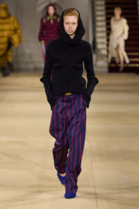 Model walking the runway in a black knit hooded sweater and blue pinstriped pants with red stripes, wearing blue glittery shoes.