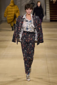 Model walking the runway in a dark floral suit with a loose patterned blouse and matching blazer draped over shoulders. Backlit, other models in background.