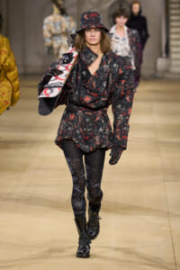 Female model walking a runway in a black and red patterned belted coat with matching hat, patterned tights, boots, and a snowboard slung over her shoulder.