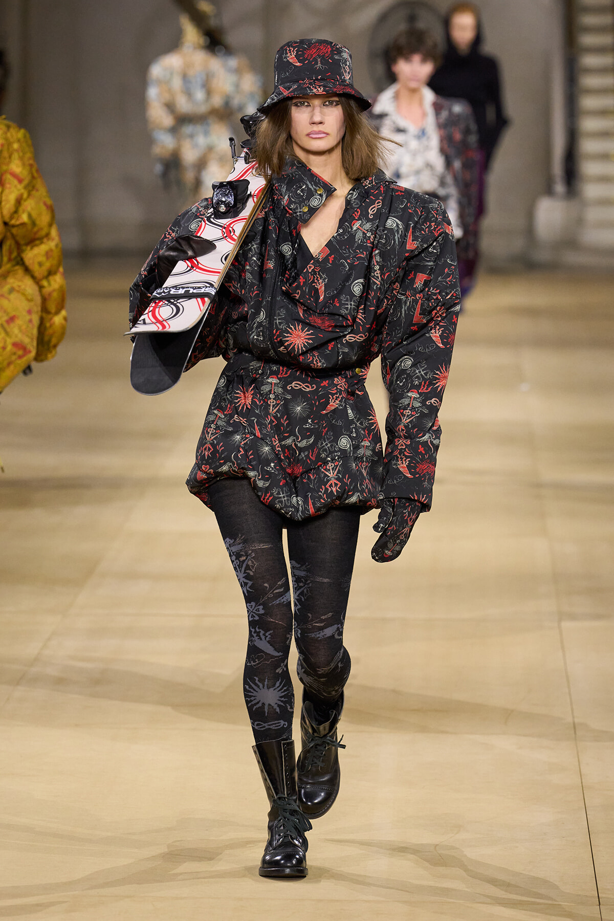Female model walking a runway in a black and red patterned belted coat with matching hat, patterned tights, boots, and a snowboard slung over her shoulder.