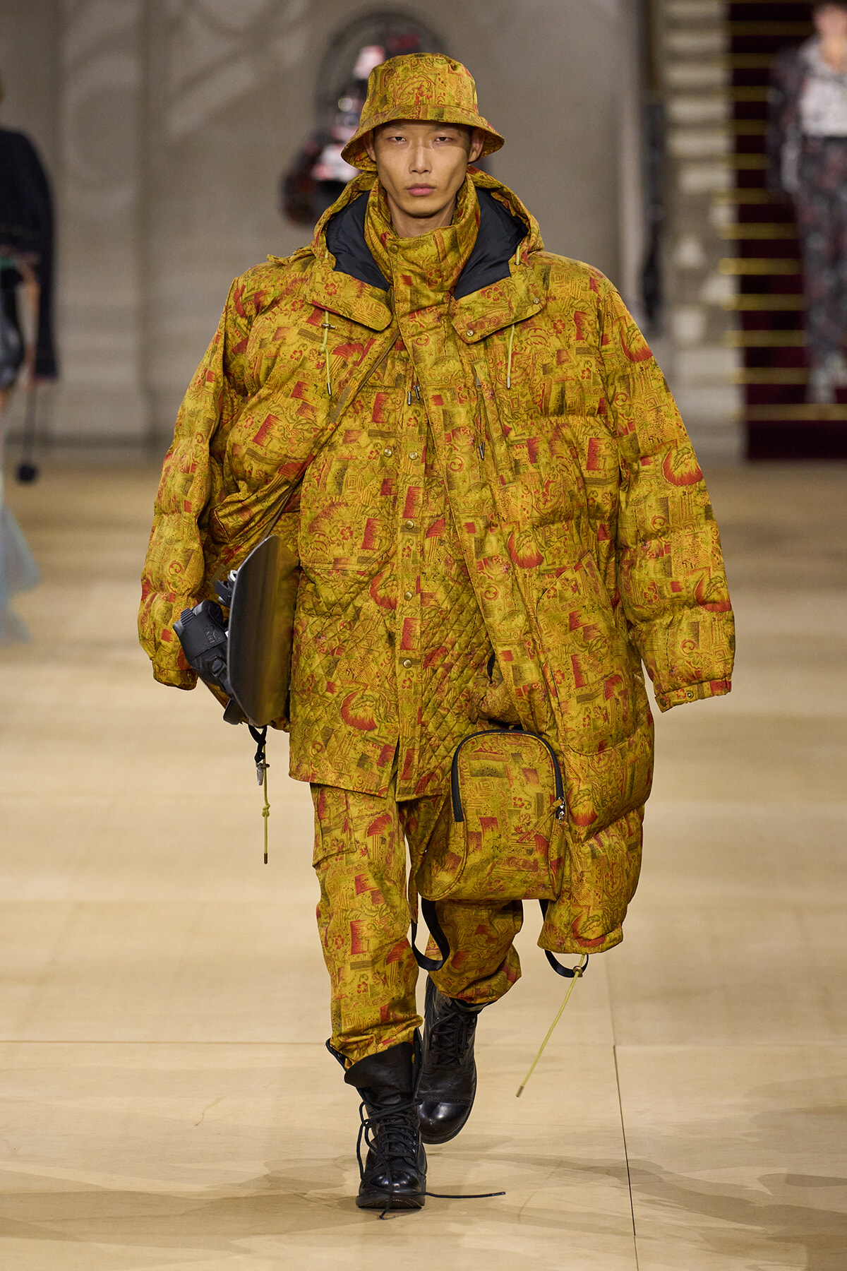 Runway model in a bright yellow patterned oversized puffer coat and matching bucket hat, carrying a black laptop bag and a yellow-accented bag.