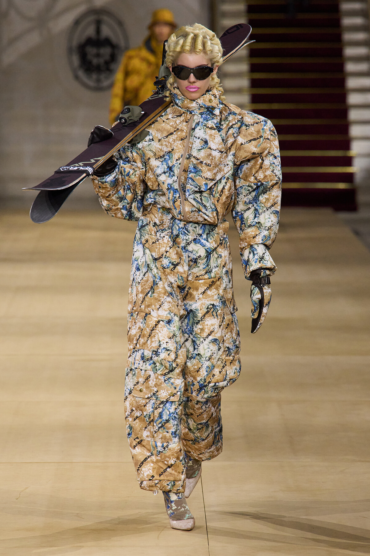 Model on a runway wearing a beige camouflage snowsuit, carrying a snowboard over her shoulder and wearing dark sunglasses.
