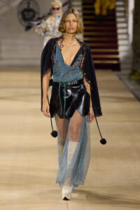 Model walks the runway in a layered outfit: teal glitter wrap over a black leather mini skirt, sheer teal overlay, and a navy cardigan with pom-pom accents, white boots.