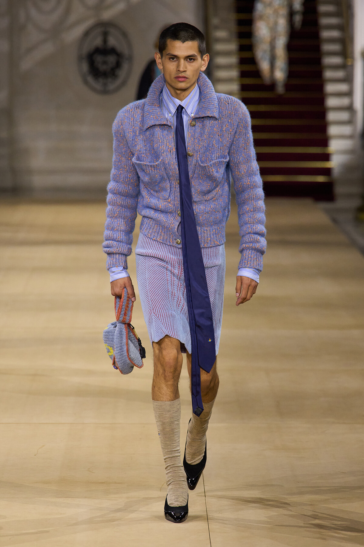 Male model walking a fashion runway in a pastel blue knit cardigan, light blue shirt, dark tie, and patterned knee-length shorts, carrying a knit bag.