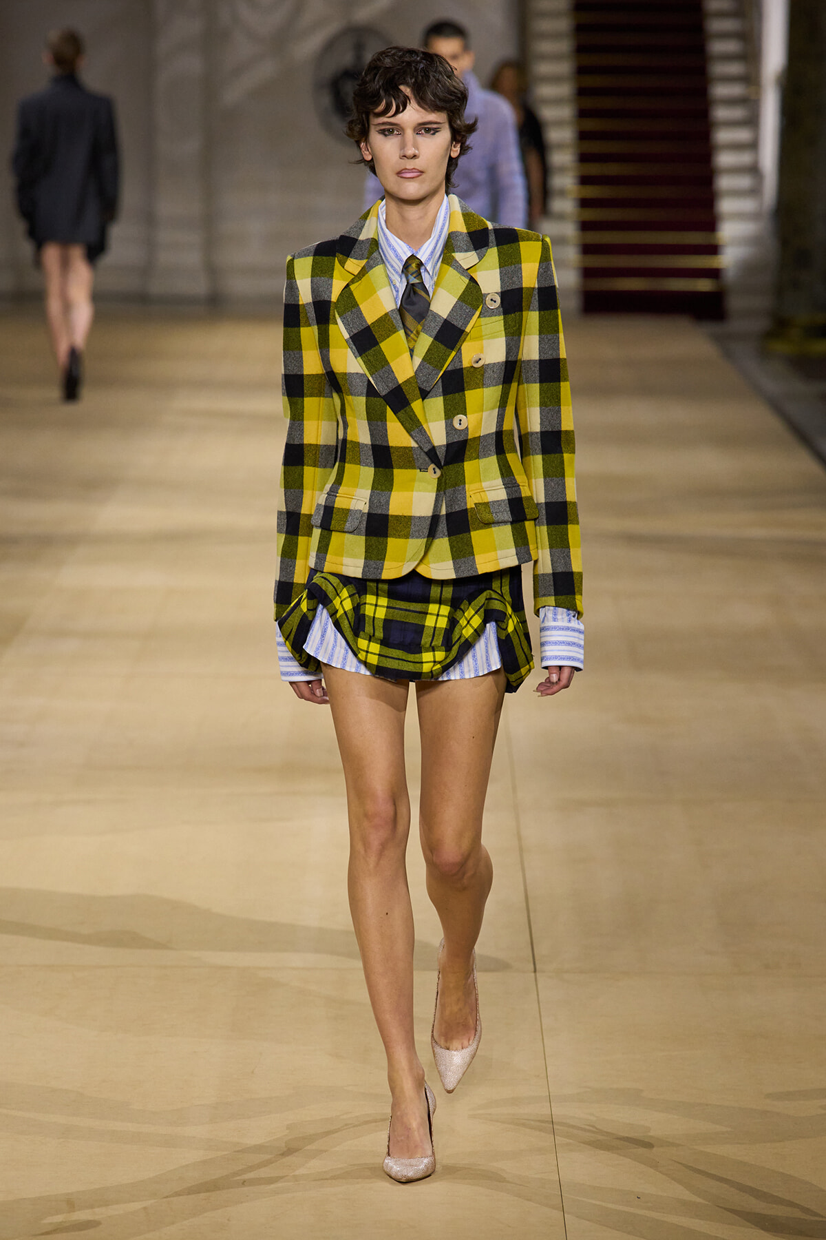 Model walks the runway in a bold yellow plaid double-breasted blazer with a matching layered plaid skirt and a blue striped shirt, nude glittery heels.