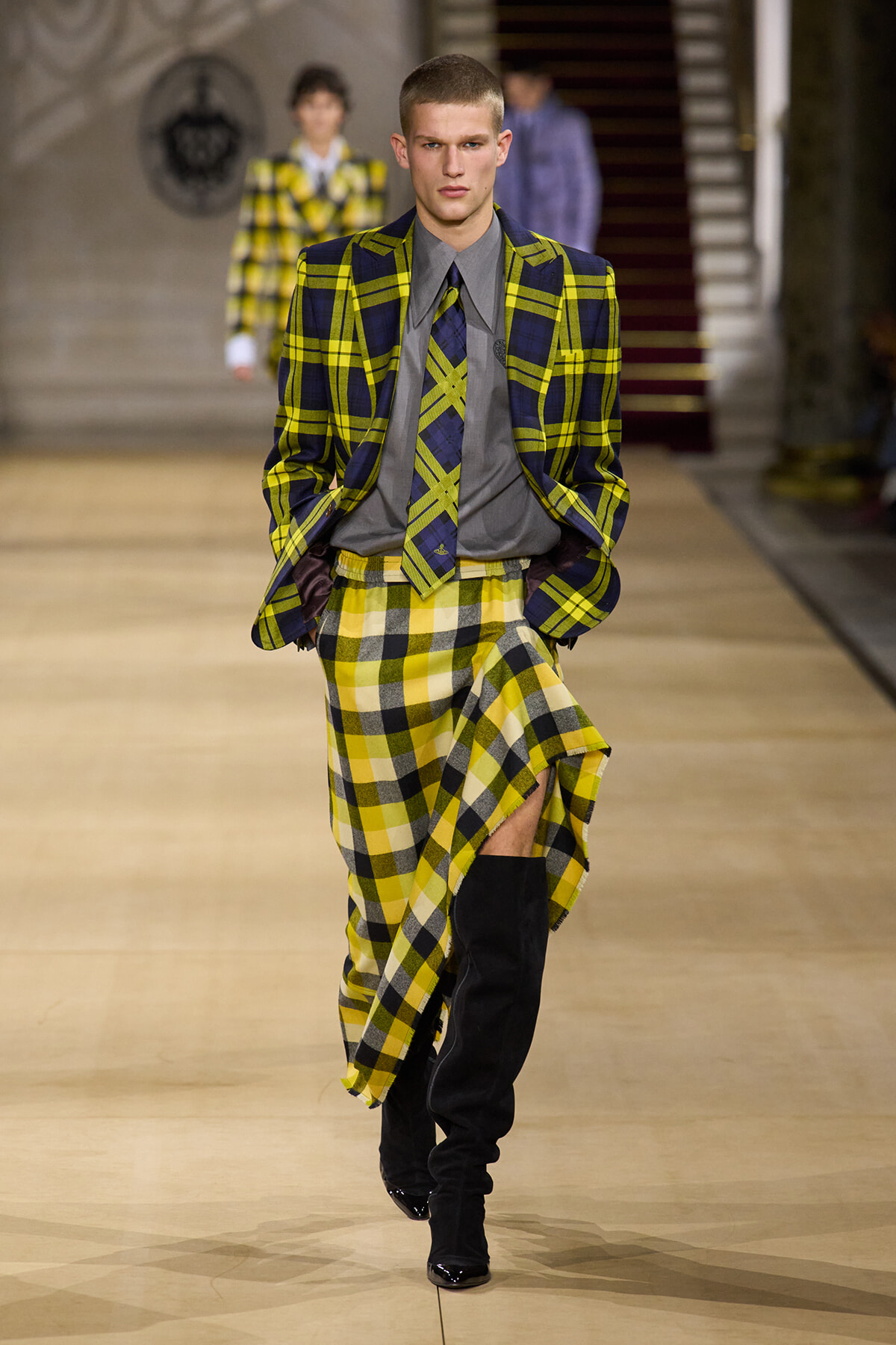 Male model walks a fashion runway in a bold yellow-and-navy plaid suit with a gray shirt and matching tie, wearing a high-low plaid skirt and black knee-high boots