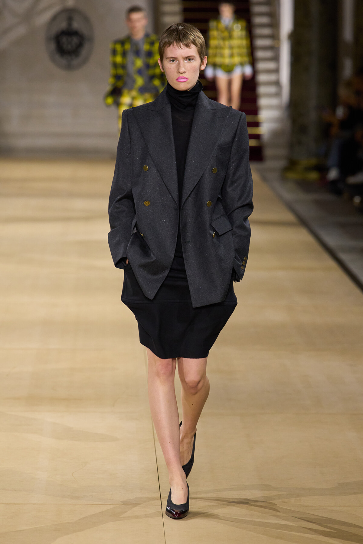Model on a runway wearing an oversized charcoal double-breasted blazer over a black turtleneck and knee-length black skirt, hands in pockets.