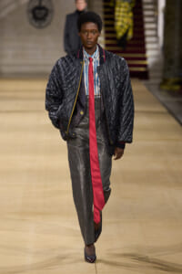 Model on a runway wearing a black quilted bomber jacket over a sparkly blazer, striped shirt, bright red tie, gray pinstripe pants, and burgundy heels.