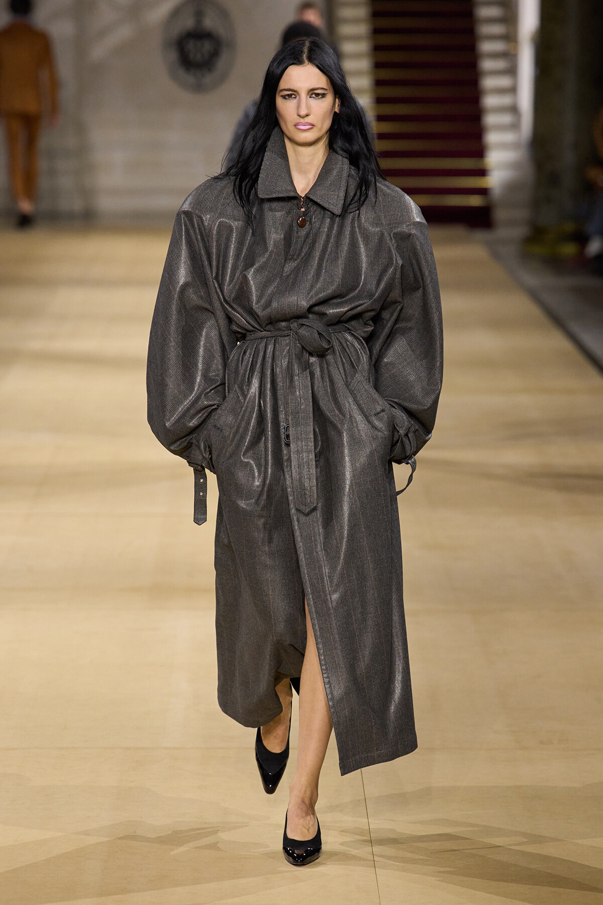 Model walks the runway in a metallic gray belted trench coat with oversized sleeves and black pumps.