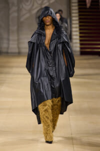 Model walks the runway in a large black shiny hooded coat over shaggy tan pants, with black heels visible at the feet.