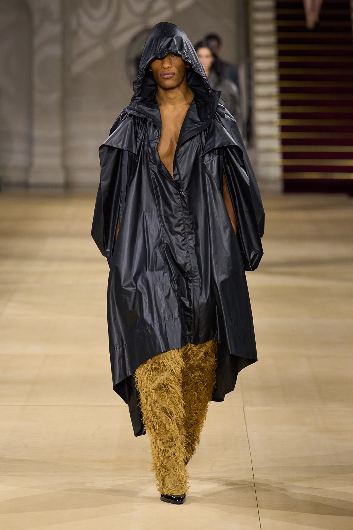 Model walks the runway in a large black shiny hooded coat over shaggy tan pants, with black heels visible at the feet.