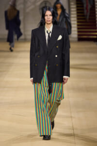 Model walks a runway wearing a dark double-breasted blazer over a light shirt with a black tie and colorful striped wide-leg pants.