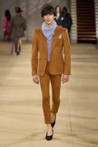 Male fashion model on a runway wearing a tan suede suit with a lavender knit scarf and matching pants, black heels, pocket square visible.