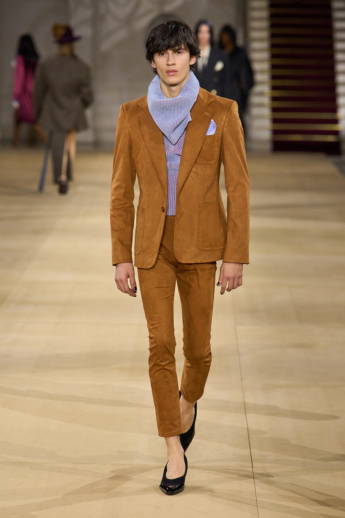 Male fashion model on a runway wearing a tan suede suit with a lavender knit scarf and matching pants, black heels, pocket square visible.